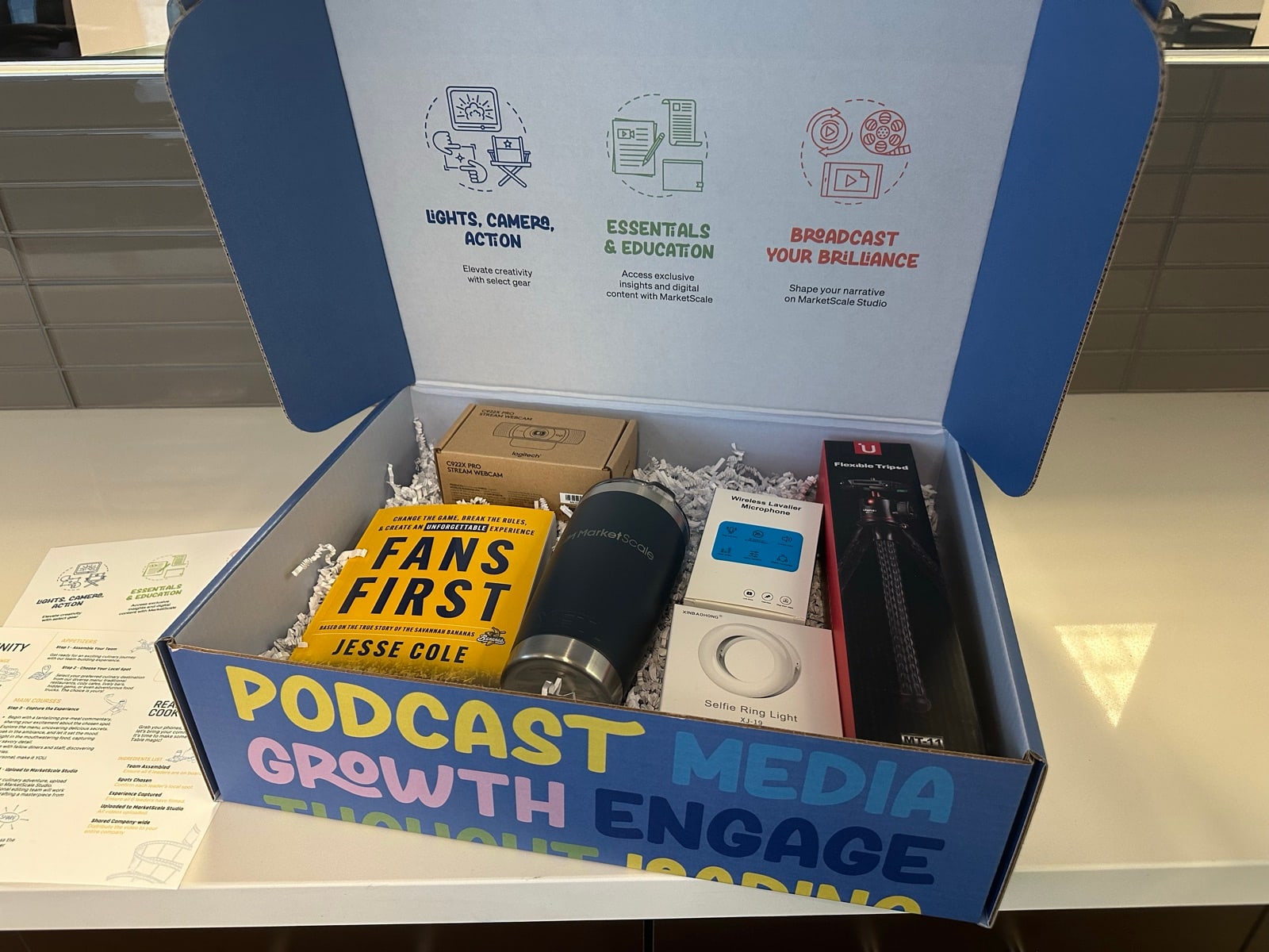 The MarketScale client kickoff package: a branded welcome box with a webcam, wireless lavalier mics, a ring light, a tripod, a MarketScale tumbler, and a welcome guide